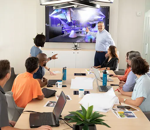Team Members sitting at a conference table looking at a speaker standing in front pointing at a screen