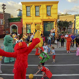 Elmo character standing with arms open on Sesame Street as children play around him